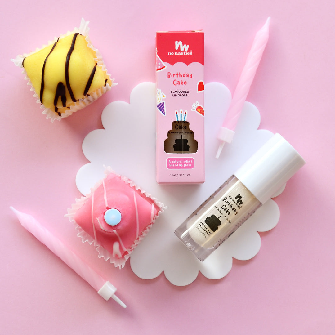 A photo of birthday cake flavored natural lip gloss for kids, presented with packaging that resembles a cupcake. The product is displayed alongside a pink wand applicator and additional cupcakes for a party theme.