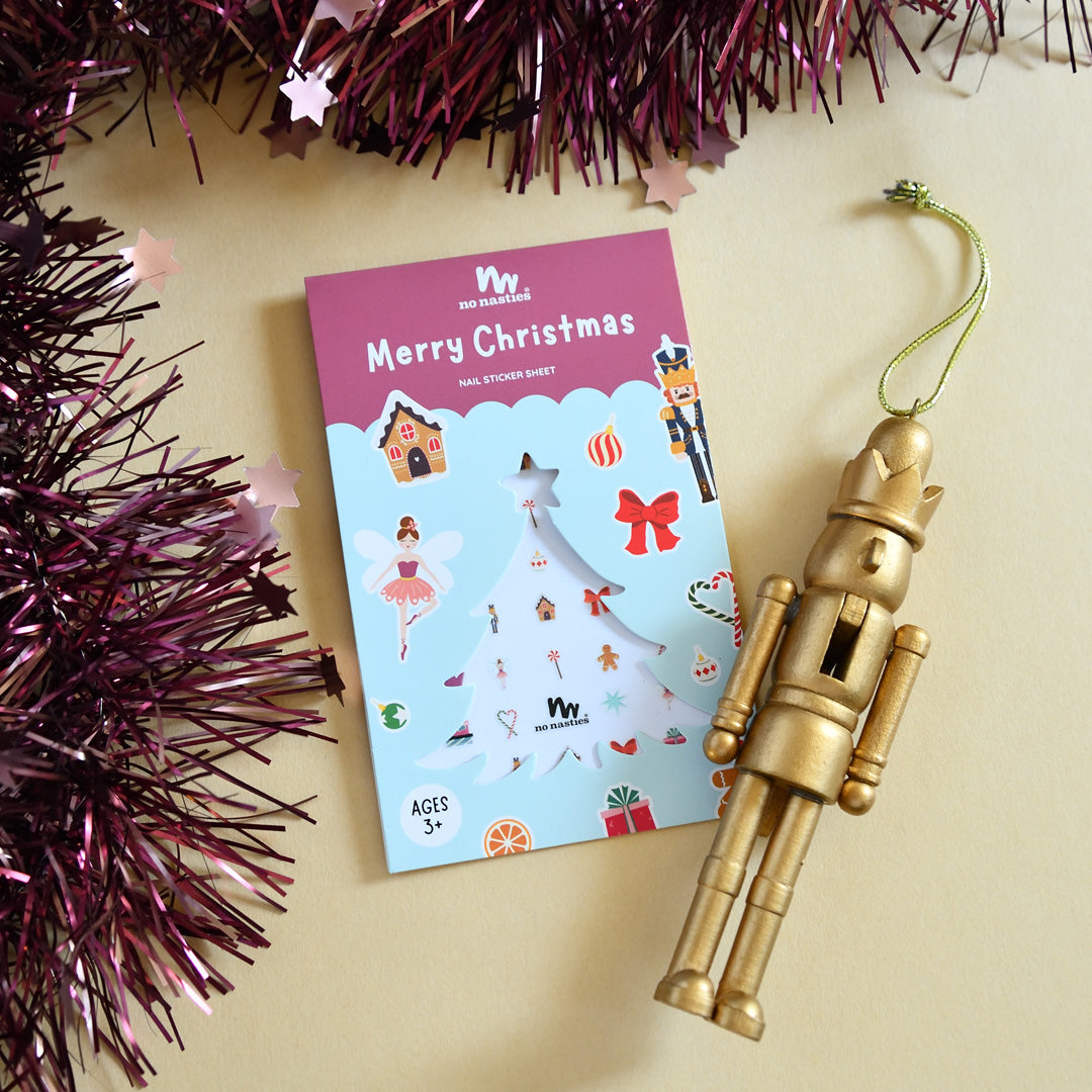A pack of Christmas themed nail stickers with various festive designs, placed next to a nutcracker Christmas ornament and with purple tinsel in the background.