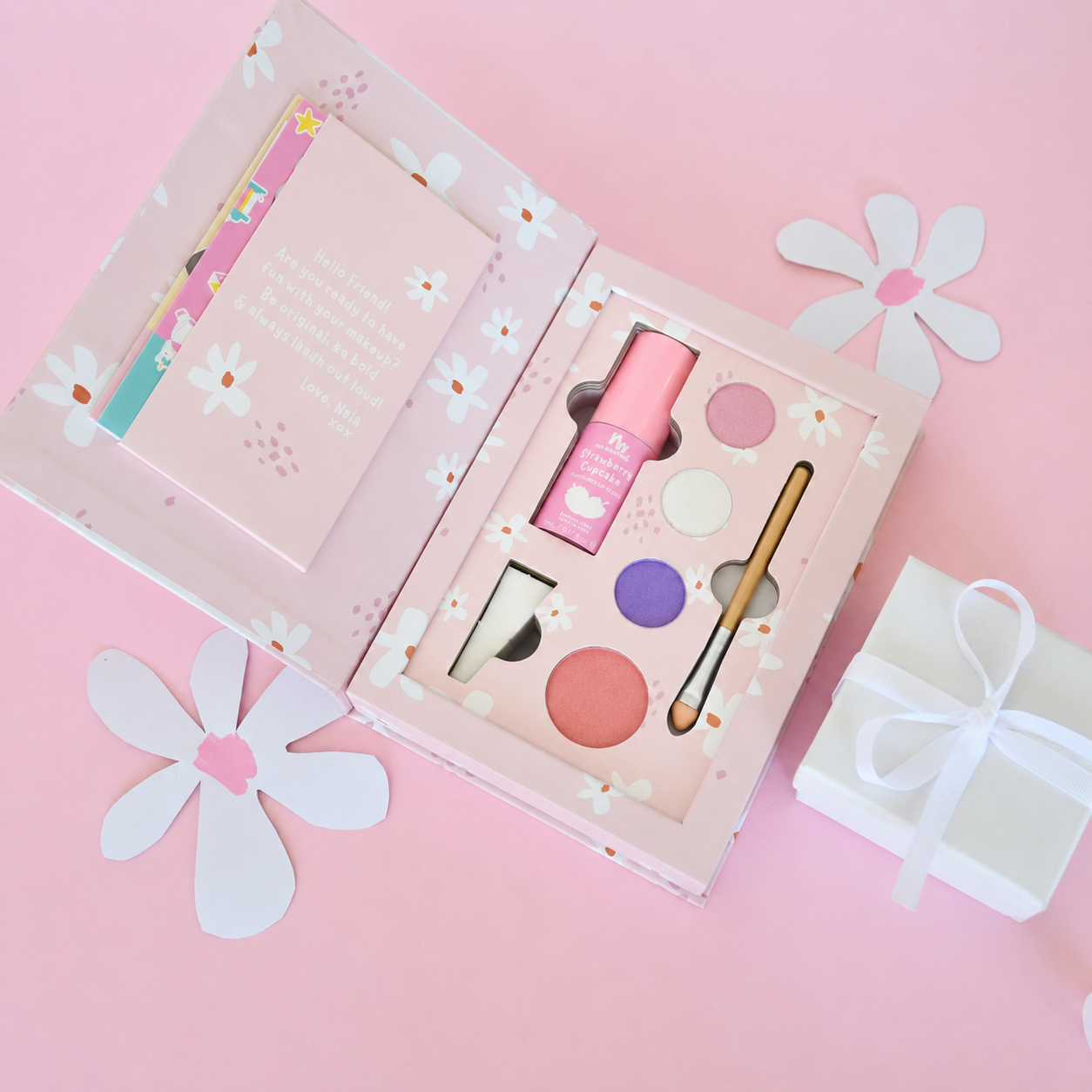 An open pink floral makeup gift set displayed on a pink background, with a small white gift box tied in ribbon beside it. The set includes a pink bottle, round eyeshadow pans in pink, purple, white, and peach, a brush, and a card inside the box.