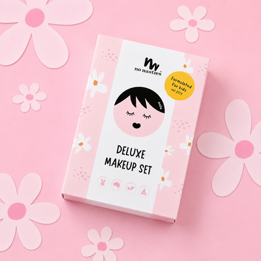 A pink and white box for the 'no nasties Deluxe Makeup Set' featuring a cute cartoon girl illustration and a yellow 'formulated for kids in 2023' badge, set against a pink background decorated with paper flowers.