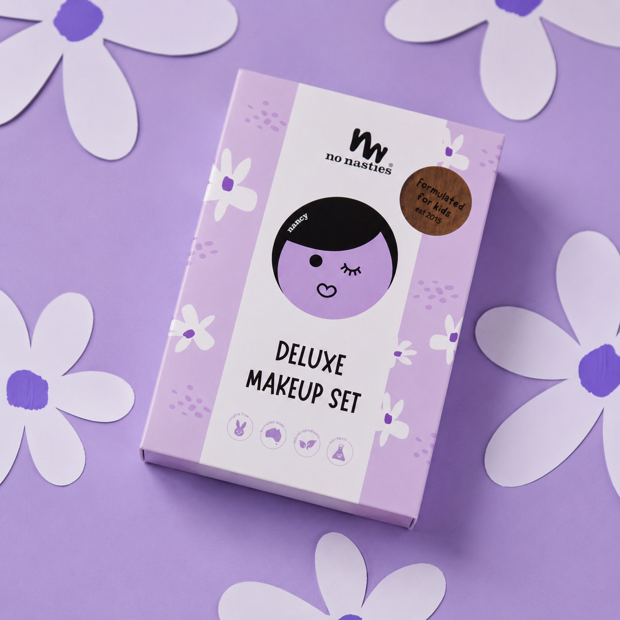 A boxed makeup set labeled 'mw no noodles DELUXE MAKEUP SET' lies on a lavender background with white paper flower cutouts around it. The box is light purple and white with a cartoon face illustration and a brown circular badge.