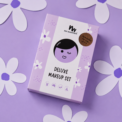 A boxed makeup set labeled 'mw no noodles DELUXE MAKEUP SET' lies on a lavender background with white paper flower cutouts around it. The box is light purple and white with a cartoon face illustration and a brown circular badge.