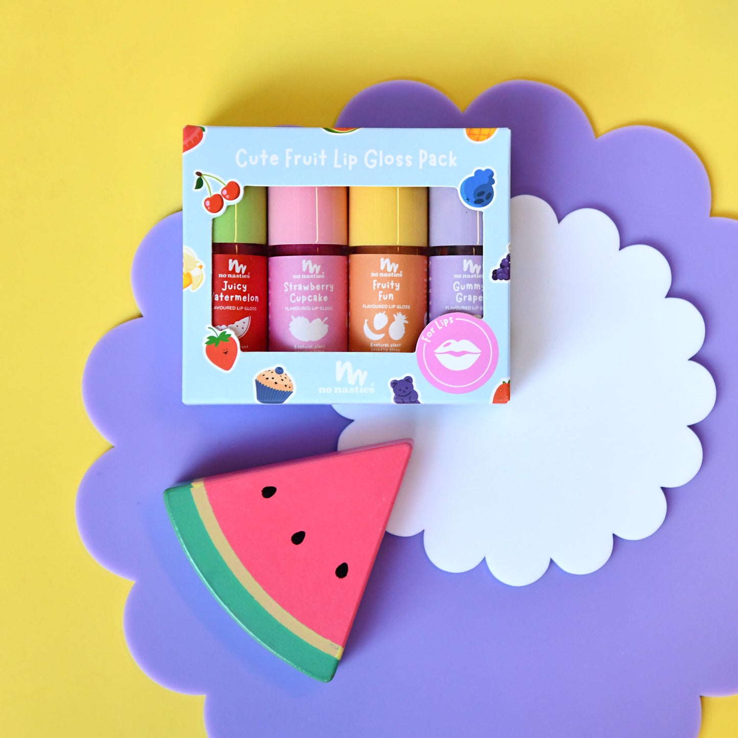 A boxed lip gloss pack labeled 'Cute Fruit Lip Gloss Pack' sits on a purple cloud-shaped prop over a yellow background. The package contains four lip gloss tubes in pink, orange, purple, and green, with a watermelon slice accessory placed in front.
