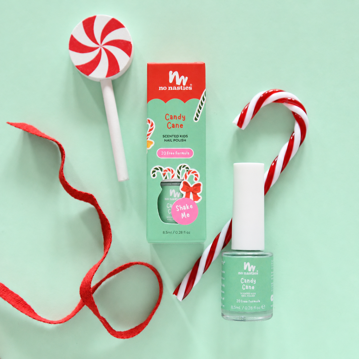 A flat-lay of a nail polish product and its box on a mint green background, styled with candy cane props and a red ribbon. The product is 'Candy Cane' by 'no nonsense' nail polish, in a mint green shade named 'Shake Me'.