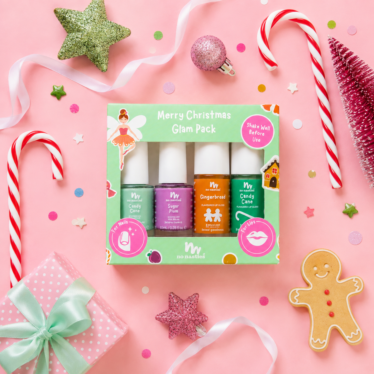A boxed nail polish set labeled 'Merry Christmas Glam Pack' arranged on a pink holiday-themed background with candy canes, glitter stars, a gift, and a gingerbread cookie. The set contains four nail polishes in green, purple, orange, and teal shades in a mint green box.