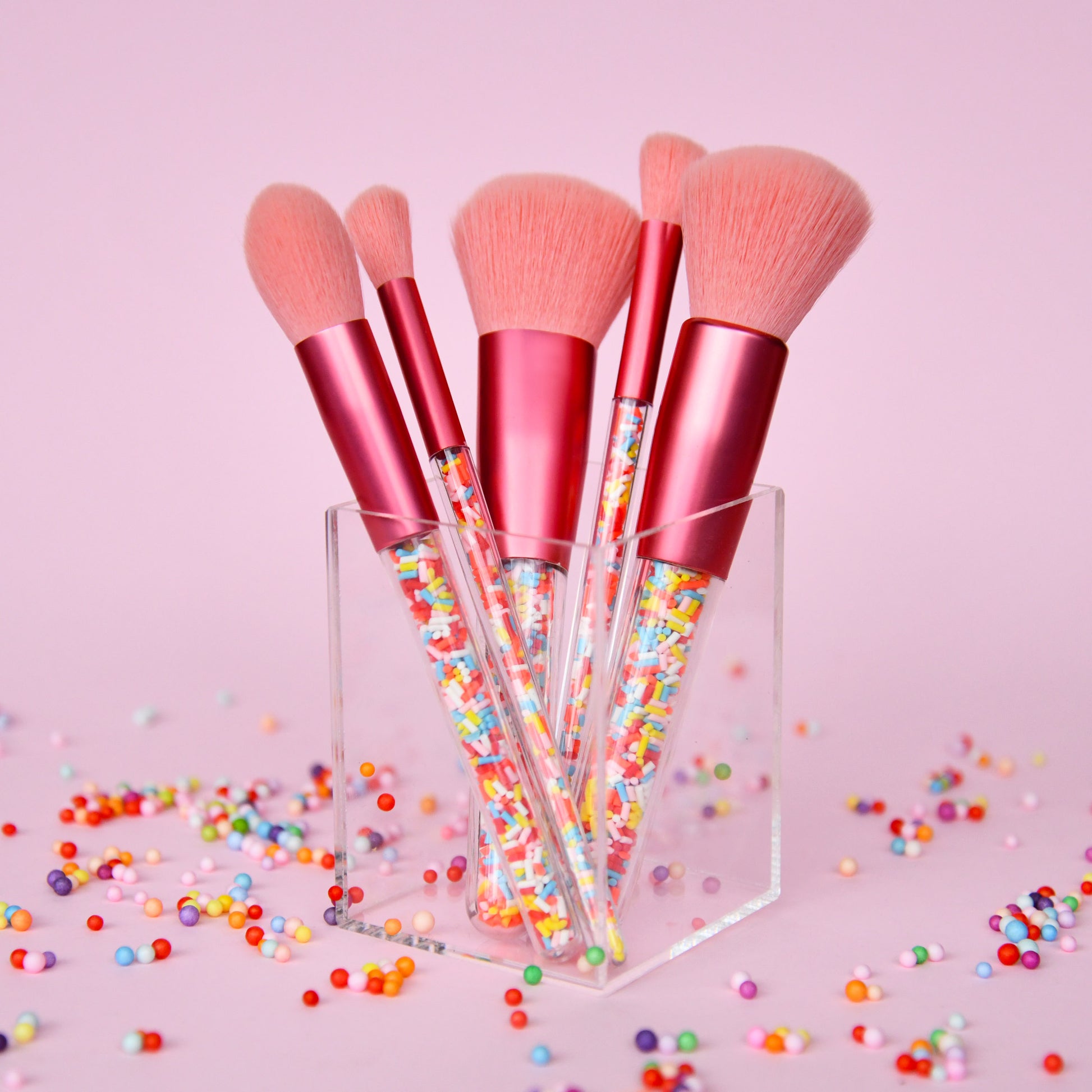 Set of makeup brushes with pink handles and clear acrylic stands on a pink background with colorful beads.