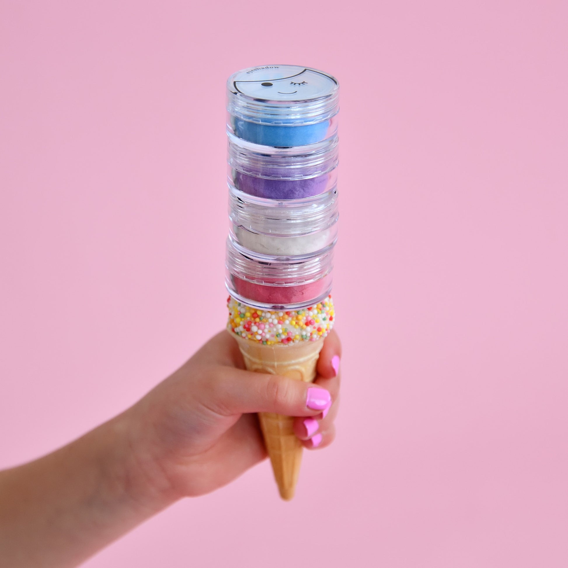 A hand holding a ice cream cone with eyeshadow colors dripping out, against a pink background.