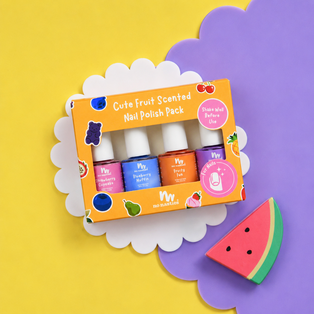 A boxed nail polish pack labeled 'Cute Fruit Scented Nail Polish Pack' by no nonsense kit, shown on a bright yellow background with purple and white scalloped paper shapes and a watermelon slice prop. The box contains four nail polish bottles in pink, blue, orange, and purple with white caps.