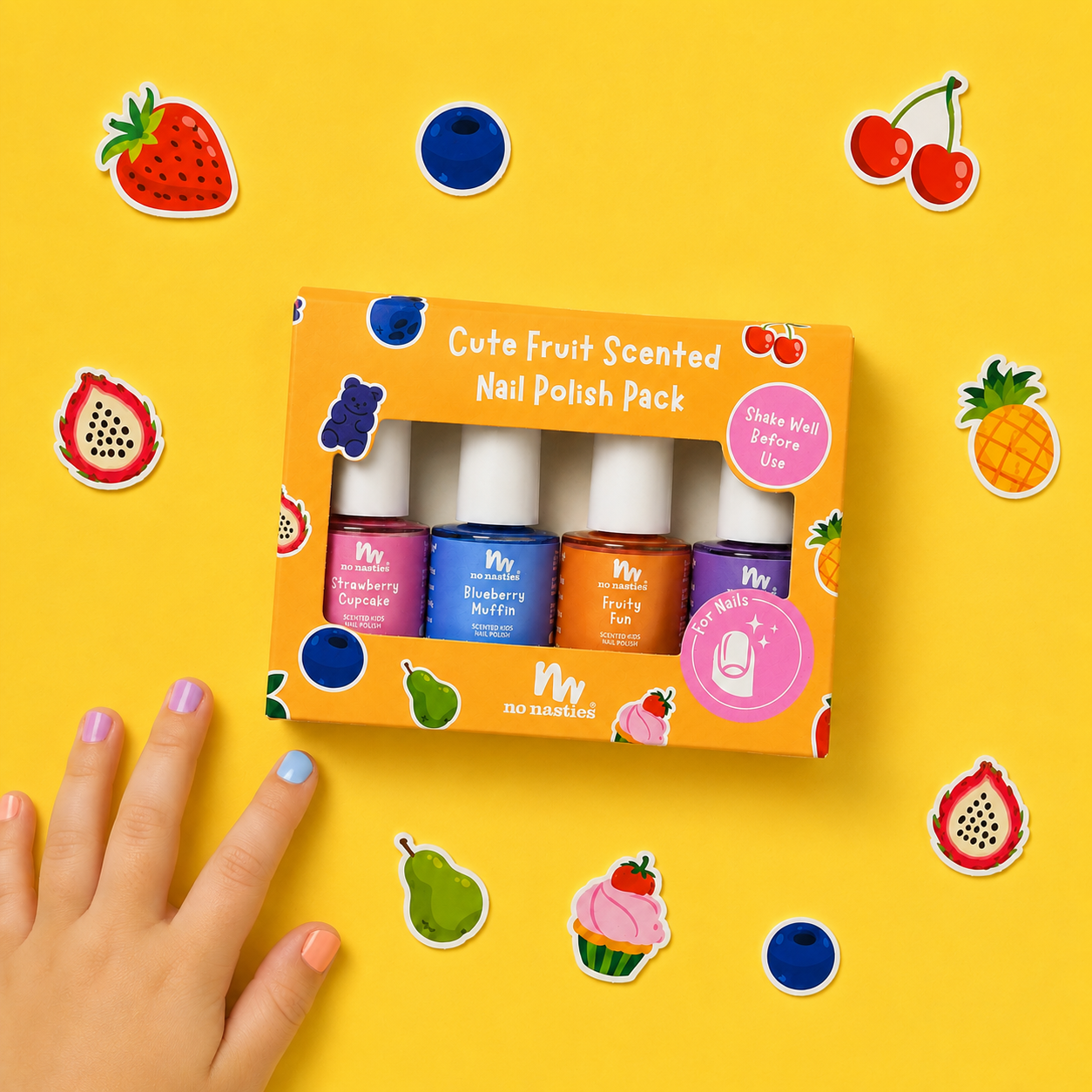 A hand with pastel-colored nail polish is shown beside a boxed set of four fruit-scented nail polishes on a bright yellow background decorated with fruit stickers. The box reads 'Cute Fruit Scented Nail Polish Pack' and shows pink, blue, orange, and purple polish bottles.