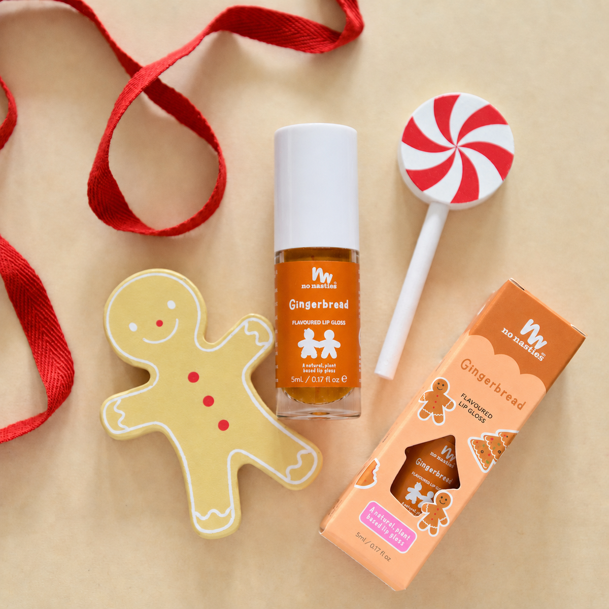 A holiday-themed nail polish set from Wiggly Sticks, featuring a Gingerbread scent. The set includes a clear bottle of orange-brown nail polish, its orange packaging box, a gingerbread man cookie-shaped lip balm, and a red and white peppermint lollipop, all arranged on a beige background with a red ribbon.