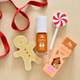 A holiday-themed nail polish set from Wiggly Sticks, featuring a Gingerbread scent. The set includes a clear bottle of orange-brown nail polish, its orange packaging box, a gingerbread man cookie-shaped lip balm, and a red and white peppermint lollipop, all arranged on a beige background with a red ribbon.