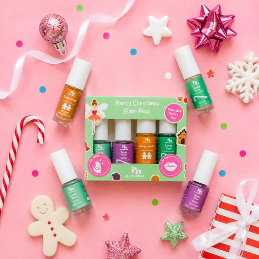A boxed nail polish set labeled 'Merry Christmas Glam Pack' surrounded by four loose nail polish bottles on a pink background with holiday props like candy canes, ribbons, and a gingerbread cookie. The set includes orange, purple, green, and peach/nude polishes.