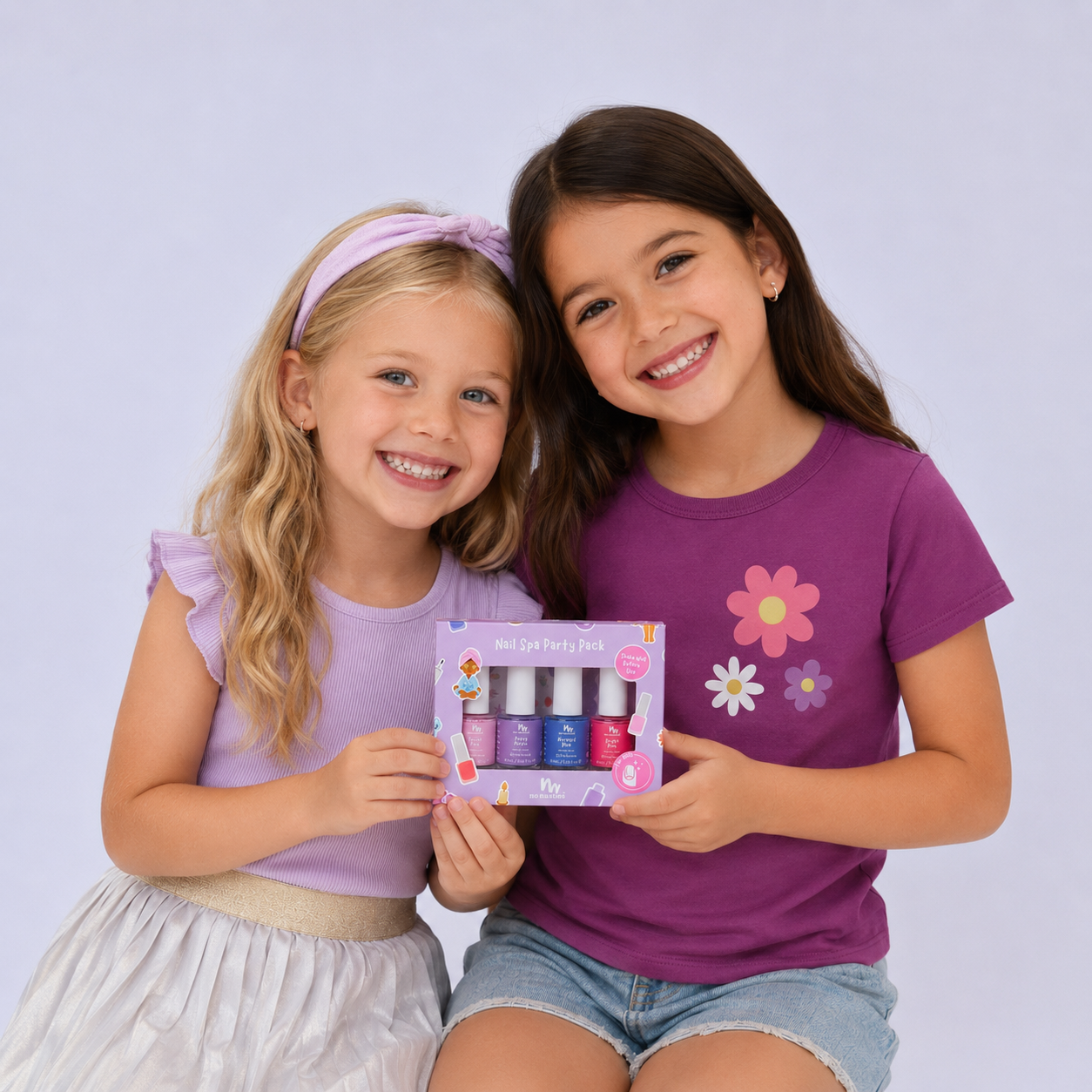 two girls sitting, holding a day spa set between them, on pastel purple background. One girl is brunette and the other blonde with a pastel purple headband and top.