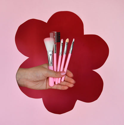 Hand holding pink makeup brushes against a red flower-shaped cutout on a pink background