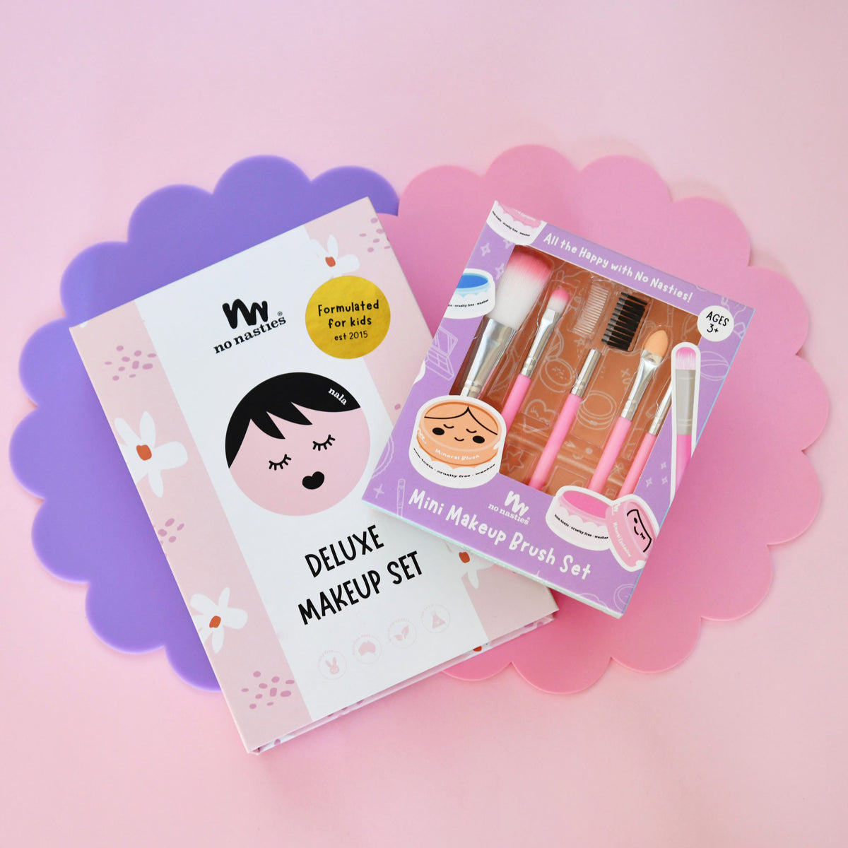 Two makeup sets, one labeled 'Deluxe Makeup Set' and the other 'Mini Makeup Brush Set', on a pink and purple scalloped background.