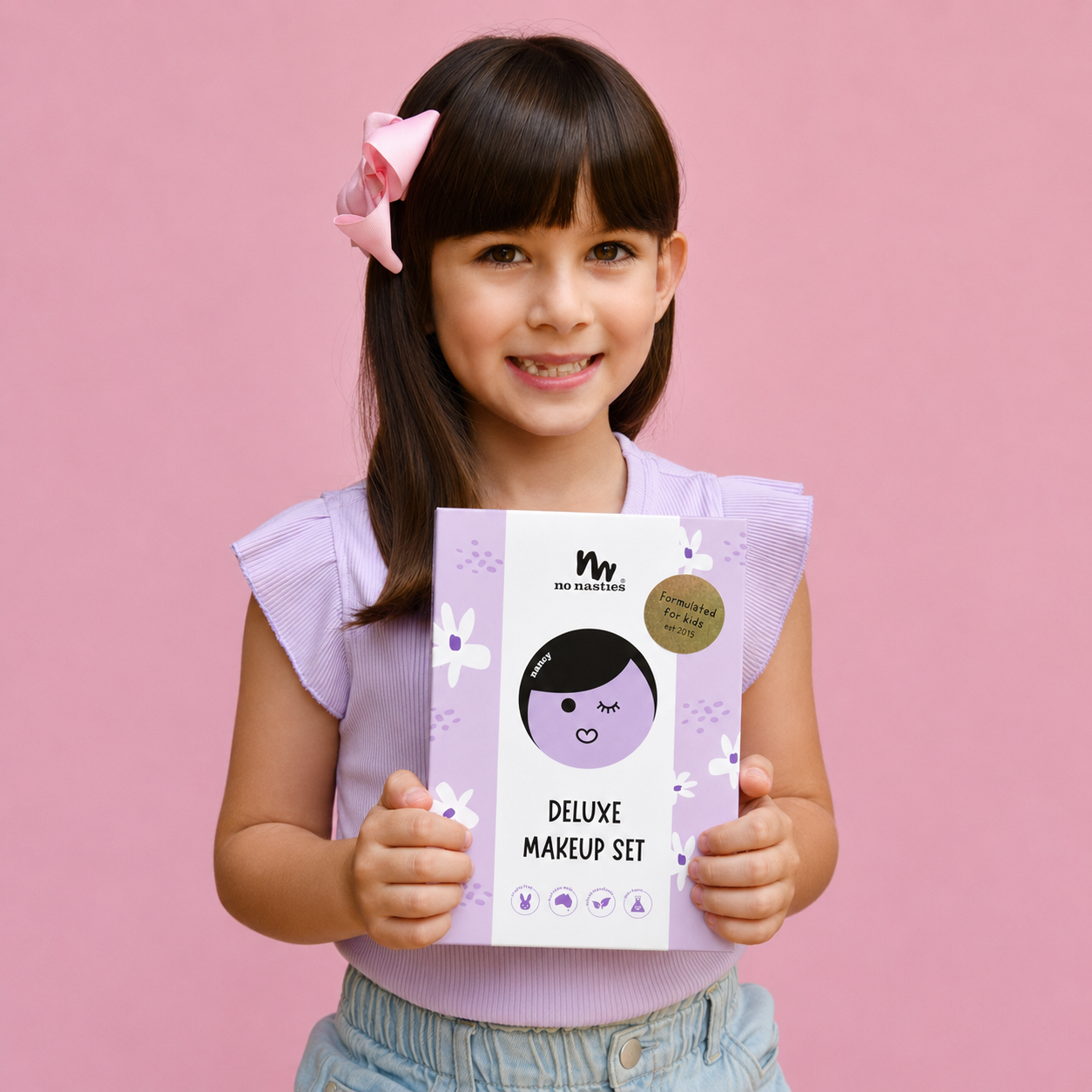 A young girl with a pink bow in her hair holds a 'Wink Magic Deluxe Makeup Set' package against a solid pink background.