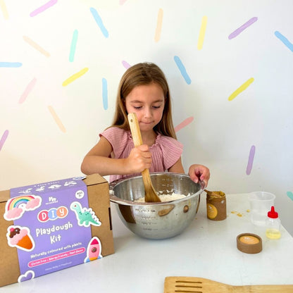 Young girl mixing playdough in a bowl with a wooden spoon, surrounded by playdough kit packaging and ingredients.