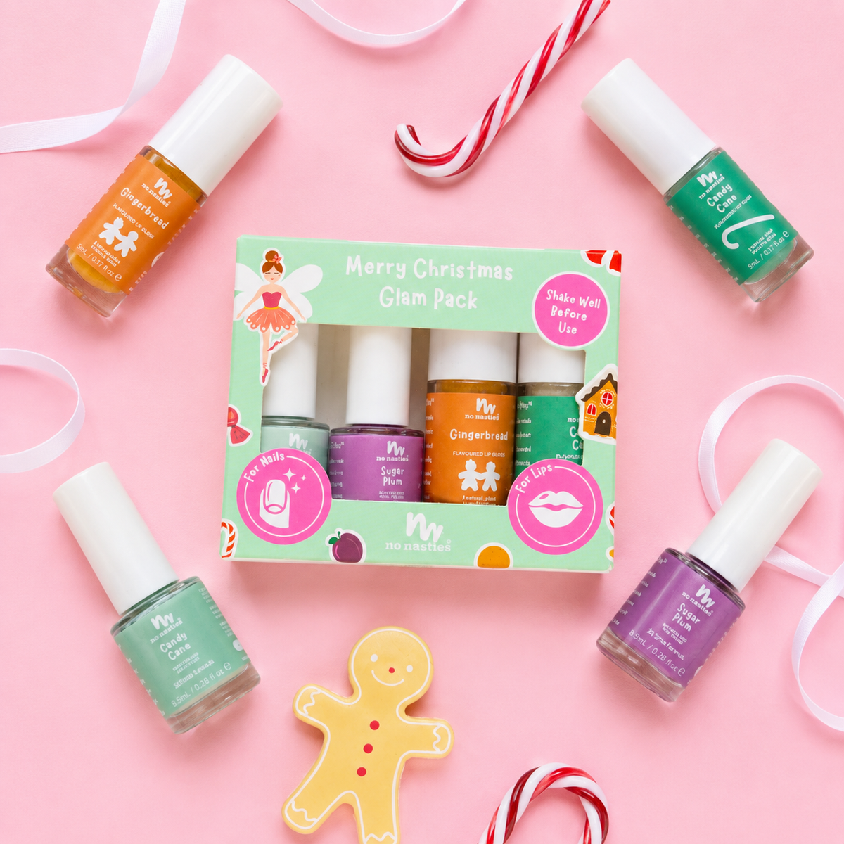 A boxed nail polish set labeled 'Merry Christmas Glam Pack' surrounded by four loose nail polish bottles on a pink background with holiday props like candy canes, ribbons, and a gingerbread cookie. The set includes orange, purple, green, and peach/nude polishes.