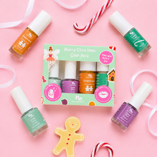 A boxed nail polish set labeled 'Merry Christmas Glam Pack' surrounded by four loose nail polish bottles on a pink background with holiday props like candy canes, ribbons, and a gingerbread cookie. The set includes orange, purple, green, and peach/nude polishes.
