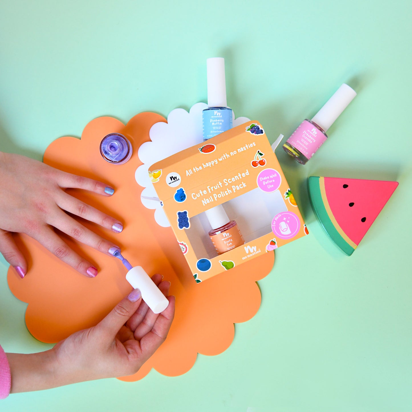 A colorful nail polish set displayed on a mint green background, featuring a hand with lavender-painted nails near an orange scalloped mat. The set includes a box labeled 'Cute Fruit Scented Nail Polish Pack', several nail polishes in blue, pink, and orange, and a watermelon-shaped item.