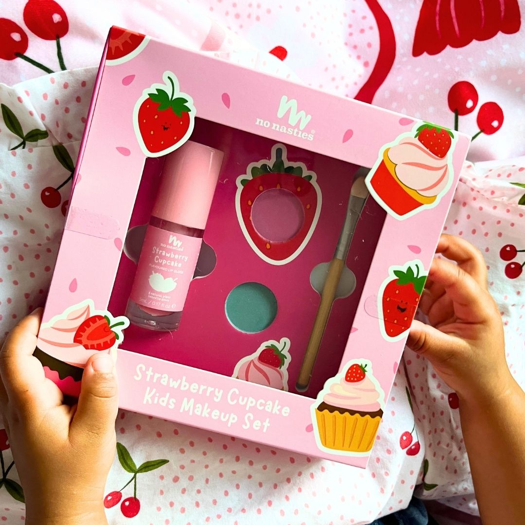 Strawberry-themed kids makeup set in a pink box held by hands on a matching background.