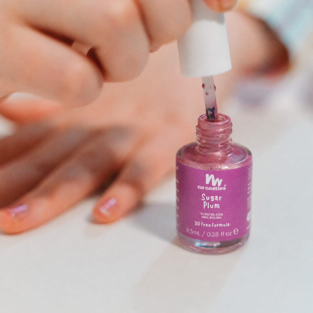 A close-up of a hand applying pink nail polish from a bottle labeled 'no nonsense Sugar Plum' onto another hand with matching pink nails, on a white surface.