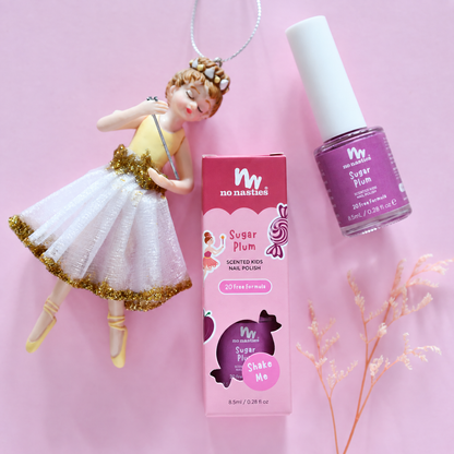 A bottle of No Nasties Sugar Plum nail polish and its pink box are arranged on a pink background next to a ballerina keychain charm.